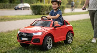 Voiture électrique enfant et homologation route : jeune enfant casqué dans une Audi rouge sur l'herbe le long d'une route où circule une vraie voiture, un parent à la télécommande