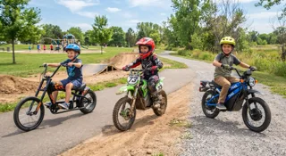 Moto électrique enfant : trois enfants côte à côte sur un chopper noir, une moto cross verte et un trail bleu sur une piste, pour comparer les trois types