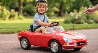 Première voiture électrique à 3 ans : jeune enfant casqué au volant d'un cabriolet rouge de style vintage dans un parc, un parent tenant la télécommande parentale