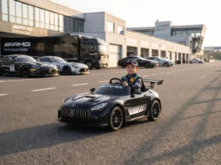 Mercedes-AMG GT3 12V noir, enfant au volant devant les stands d'un circuit, voitures de course en arrière-plan